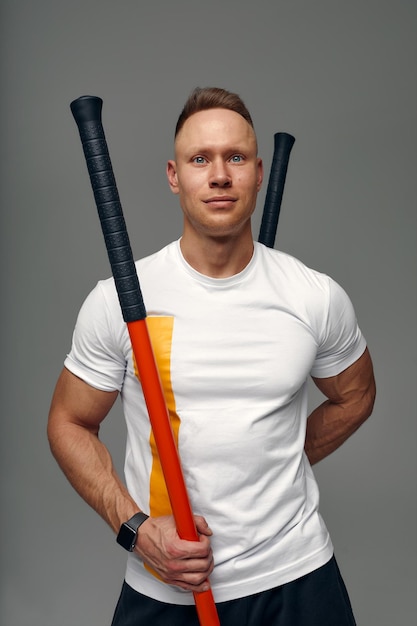 Un combattant en t-shirt blanc et short noir se tient dans une pose artificielle de combat et tient un bâton de combat sur fond de studio