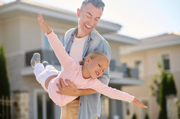 Comme un avion. Papa heureux tenant sa petite fille blonde inspirée jouant un avion dans la rue par une belle journée chaude