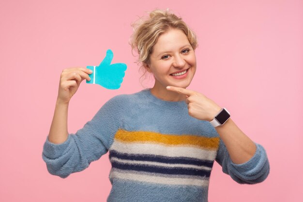 Comme le bouton. Portrait d'une femme joyeuse en chandail pointant vers l'icône bleue du pouce vers le haut, notification des abonnés, recommandation et bons commentaires dans les médias sociaux. tourné en studio isolé sur fond rose