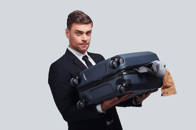 Comment gérer ces bagages ? Beau jeune homme portant une valise pleine sur tout en se tenant debout sur fond gris