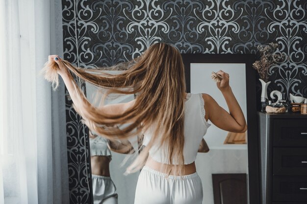 Comment prendre soin des cheveux longs vue arrière d'une jeune femme blonde naturelle en vêtements de maison blancs la peignant