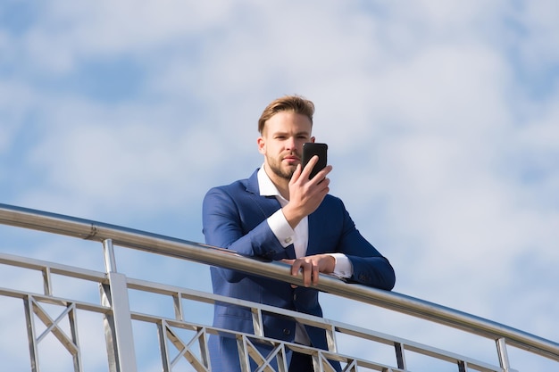 Communication d'entreprise, nouvelles technologies, sms. Homme avec smartphone sur terrasse sur ciel bleu, communication d'entreprise. Homme d'affaires en costume d'affaires avec téléphone portable, nouvelle technologie. Vie moderne, style de vie