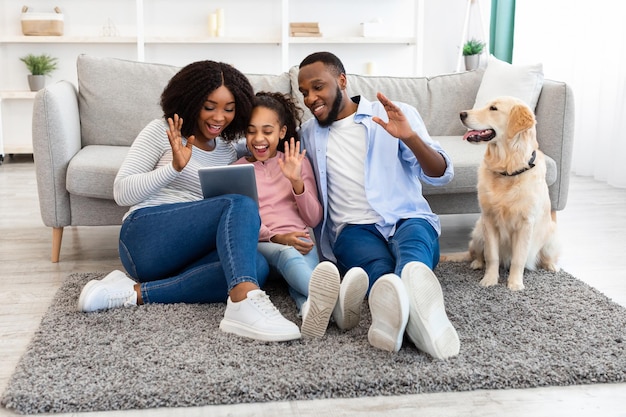 Communication en ligne. Homme afro-américain souriant, femme et fille faisant un chat vidéo numérique avec des amis ou la famille à l'aide d'une tablette, saluant la webcam, assis avec un chien sur le tapis du sol dans le salon