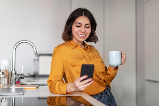 Communication en ligne Jeune femme arabe utilisant un smartphone et buvant du café dans la cuisine