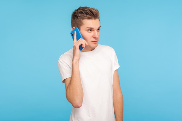 Communication mobile. Portrait d'un homme en t-shirt blanc décontracté faisant appel à un partenaire à consulter, ayant une conversation sérieuse sur un téléphone portable, discutant de mauvaises nouvelles. studio d'intérieur tourné isolé sur fond bleu