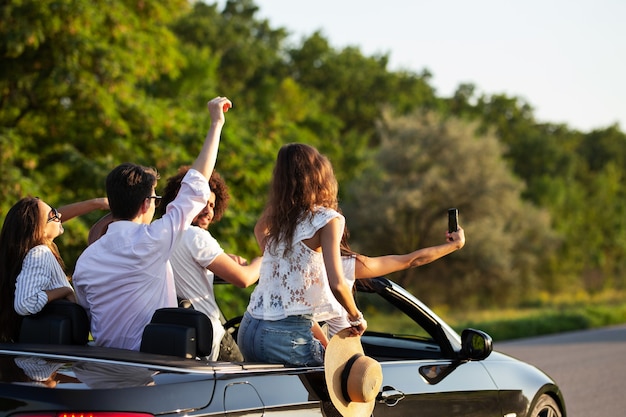 Une compagnie de jeunes femmes et d'hommes fait un selfie dans un cabriolet noir sur la route par une chaude journée ensoleillée. .