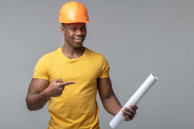 Composant important. Homme optimiste joyeux dans le casque protecteur et le tshirt jaune pointant avec le doigt au rouleau de papier