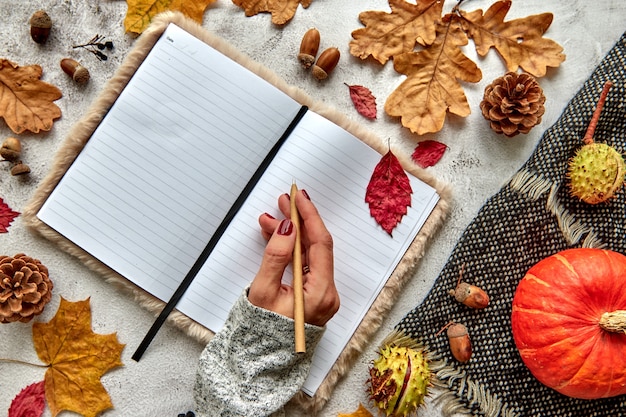 Composition d'automne, d'automne ou d'halloween faite de feuilles séchées, citrouille, pommes de pin, glands, écharpe chaude et main avec un stylo sur fond de béton. Cahier vierge de maquette de modèle avec espace de copie.