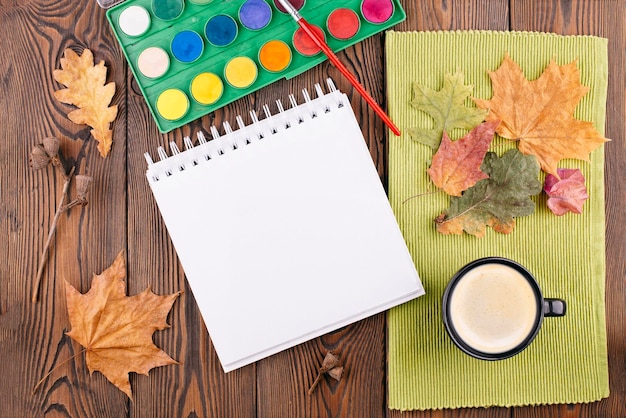 Composition d'automne avec un carnet de croquis vide avec un ressort une palette d'aquarelles automne feuilles sèches une tasse de café sur un fond en bois Espace de copie plat