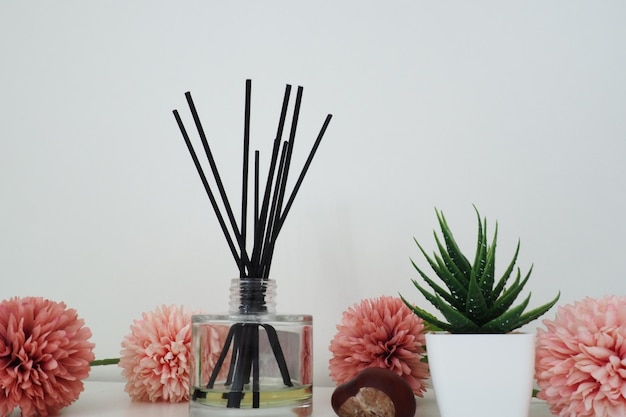 Composition de bâtons d'encens de fleurs artificielles roses dans un vase en verre avec de l'huile essentielle Nature morte sur une table blanche Fleur d'aloès dans un pot blanc Décoration de chambre et d'appartement Design d'intérieur