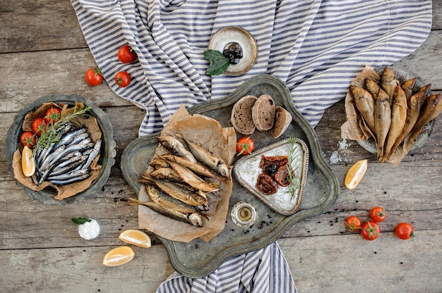 Composition délicieuse sur la table en bois et serviette à rayures grises du chinchard séché à la fumée et des anchois avec tomates, romarin, citron, pain et autres produits savoureux.