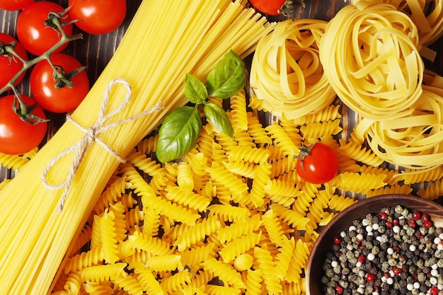 Composition avec différentes pâtes non cuites, ingrédients pour la sauce. tomates cerises, épices, huile, basilic sur fond de bois. vue de dessus. Place pour le texte.