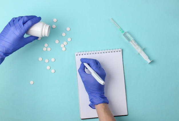 Composition du lieu de travail de médecin aboyant plat. Les mains d'un médecin avec des gants écrivent dans le bloc-notes, les pilules, la seringue sur un bleu. Vaccination épidémique