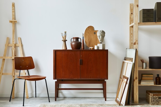 Composition élégante de l'espace de travail de l'artiste avec commode rétro en teck, chaise, bibliothèque, cadres, chevalet, décoration et accessoires personnels élégants.