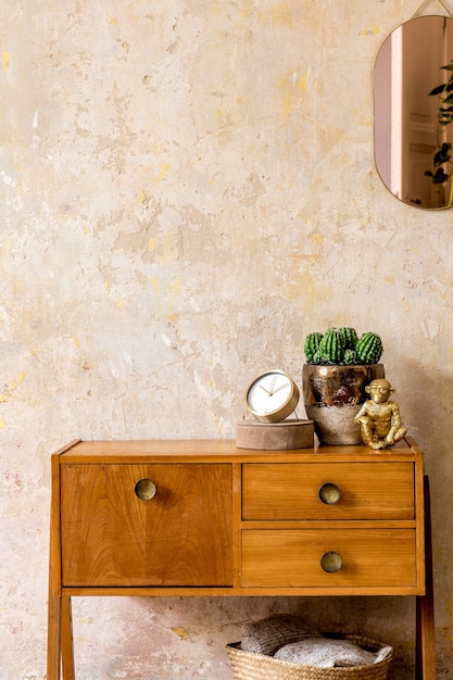 Composition élégante de l'intérieur du salon rétro avec commode vintage en bois, miroir rose doré, plante, panier en rotin, plaid, décoration et accessoires personnels élégants dans le décor de la maison wabi sabi.