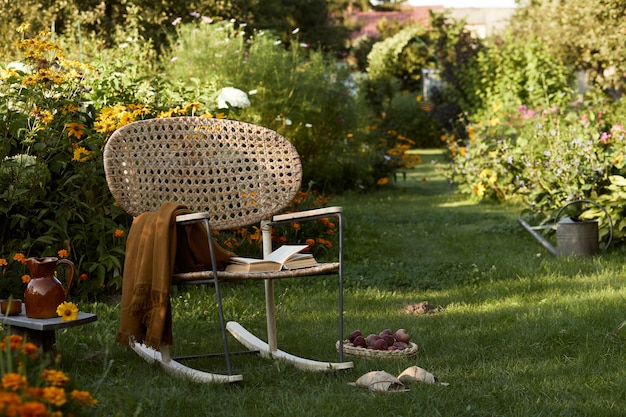 Composition élégante de jardin de campagne avec fauteuil design en rotin, banc en bois, plaid, nourriture, boissons et accessoires élégants. Beaucoup de fleurs colorées. Ambiance estivale.