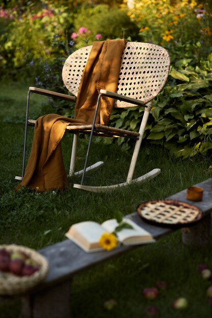 Composition élégante de jardin de campagne avec fauteuil design en rotin, banc en bois, plaid, nourriture, boissons et accessoires élégants. Beaucoup de fleurs colorées. Ambiance estivale.