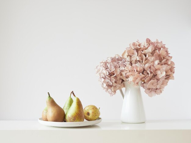 Composition élégante et simple sur une table blanche avec des fleurs d'hortensia roses séchées et un plat avec des poires Vue de face avec espace de copie sur fond de mur blanc cassé