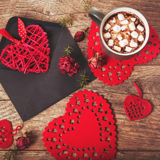 Composition festive sur table en bois rustique, mug rouge et gris avec guimauves ou cacao, coeurs dans des enveloppes, fleurs roses séchées. Copiez l'espace. Anniversaire, mère, Saint Valentin, femme, concept de jour de mariage