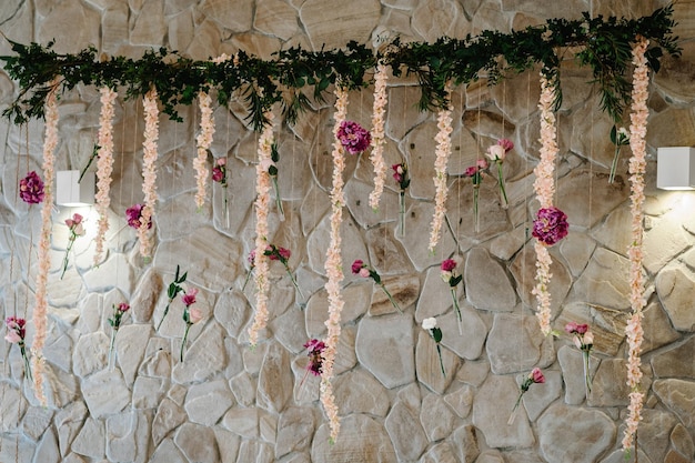 Composition d'herbes de verdure de fleurs roses violettes dans des vases en verre et des ampoules suspendues à une cérémonie de mariage sur le stand de la zone photo du mur de pierre au-dessus de la table de fête des jeunes mariés Cérémonie de mariage Arch