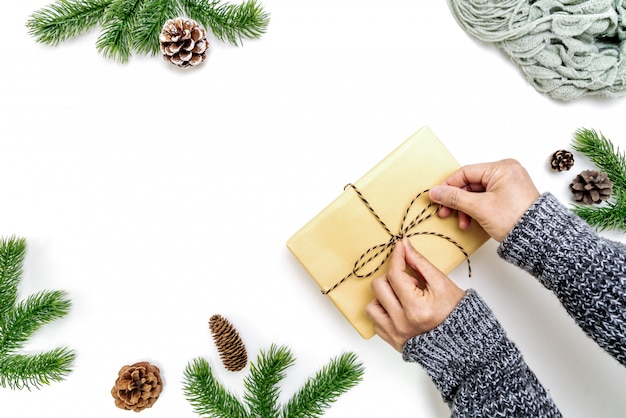 Composition d'hiver de Noël. Mains de femme enveloppant la boîte de cadeaux de Noël avec des pommes de pin, des branches de sapin sur fond blanc. Mise à plat, vue de dessus, espace copie