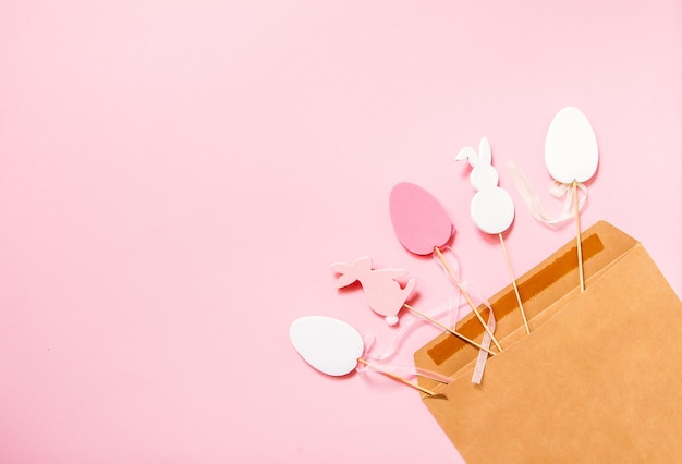 Composition horizontale de Pâques avec décor traditionnel. Oeufs colorés décoratifs et figurines en bois de lapin dans une enveloppe en papier craft sur fond de papier rose clair.