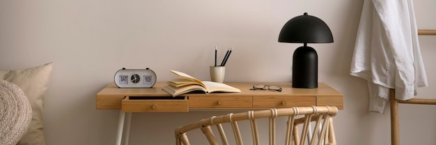 Composition minimaliste d'une chambre confortable avec espace de copie bureau en bois fauteuil boho oreillers beiges lampe noire horloge livre et accessoires personnels Modèle de décoration