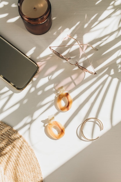 Composition de mode avec des accessoires féminins sur un tableau blanc avec une ombre de feuille. Boucles d'oreilles, bracelet, lunettes de soleil, chapeau de paille sur blanc