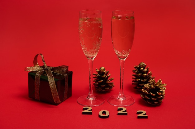 Une composition d'objet du Nouvel An, des chiffres en bois de 2022, des pommes de pin dorées, un cadeau de Noël dans du papier d'emballage pailleté avec un arc doré, des flûtes à champagne avec du vin mousseux sur fond rouge, un espace pour copie