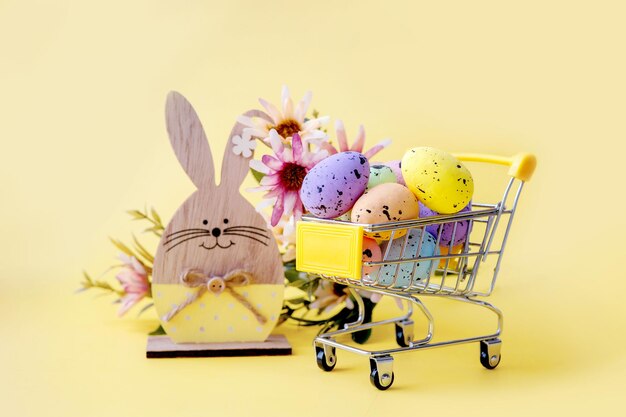 Composition de Pâques avec des œufs colorés dans un panier, un lapin en bois et des fleurs printanières sur fond jaune.