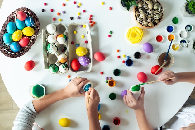 Composition de Pâques avec des peintures colorées et des œufs se trouvent sur la table. Vue de dessus les mains des enfants peignent des œufs