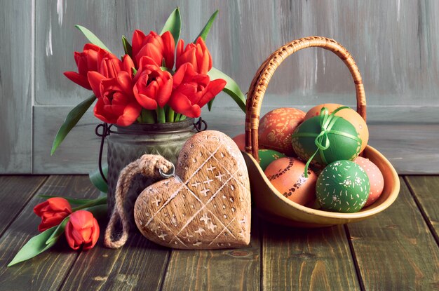 Composition de Pâques avec des tulipes rouges, un cœur en bois et un panier d'oeufs de Pâques colorés sur bois