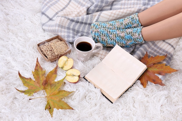 Composition avec plaid chaud, livre, tasse de boisson chaude et jambes féminines, sur fond de tapis de couleur