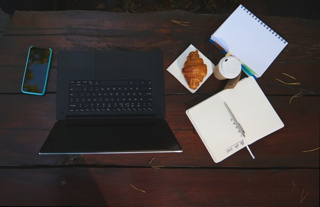 Composition à plat d'articles d'affaires et de bureau. Ordinateur portable, smartphone, cahier ouvert, bloc-notes et croissant frais avec tasse en papier à emporter de café chaud sur une surface de table en bois. Vue grand angle