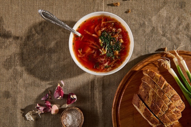 Composition à plat avec un bol en céramique avec du bortsch, des oignons verts et du pain de grains entiers tranché avec des croustilles sur une planche à découper en bois, de l'ail pelé sur une nappe en lin. Cuisine traditionnelle ukrainienne