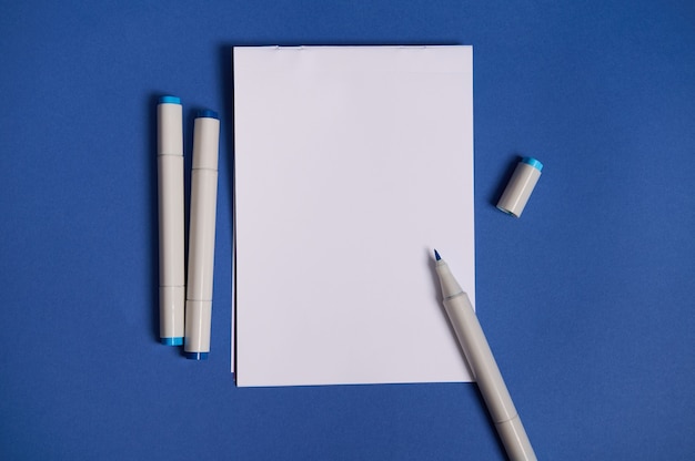 Composition à plat dichromatique avec des marqueurs aquarelles ou des feutres avec une feuille de papier vide blanche avec espace de copie, isolée sur fond bleu.