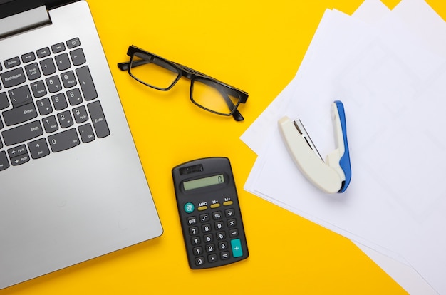 Composition à plat sur un jaune. Espace de travail de bureau avec ordinateur portable, calculatrice, lunettes, agrafeuse et papier