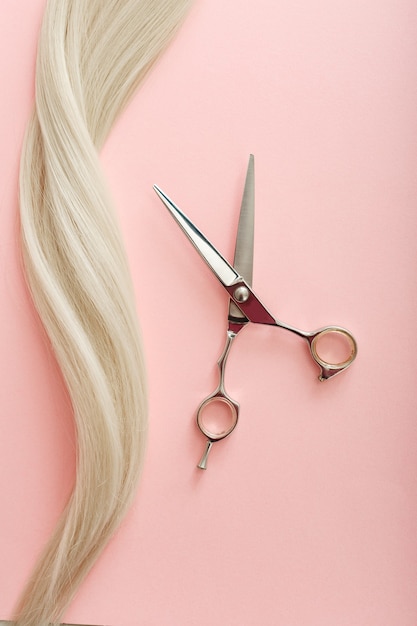 Composition à plat avec outils de coiffeur - ciseaux et mèche de cheveux blonds sur fond de couleur rose avec espace de copie pour le texte. Service de coiffeur. Prestation de salon de beauté.