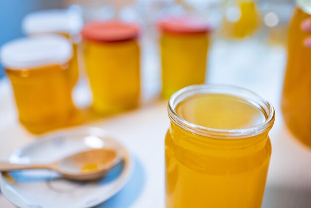 Composition de plusieurs pots transparents en verre avec du miel jaune sucré et une petite soucoupe avec une grande cuillère en bois sur une grande table blanche par une belle journée ensoleillée