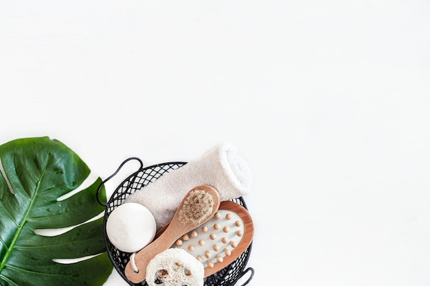 Composition de spa avec brosse de massage et autres accessoires de bain dans le panier sur un mur blanc avec feuille de monstera.