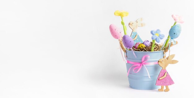 Composition de vacances pour enfants intérieurs avec petit seau bleu avec nid d'oeufs de caille colorés oeufs décoratifs fleurs de couleur tricotées lapins en bois sur bannière de Pâques blanche Carte de fête Espace de copie