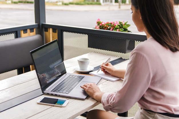 Concentré au travail. Jolie jeune femme d'affaires, pigiste ou étudiant travaillant sur ordinateur portable et prendre des notes dans son cahier à terrasse au café