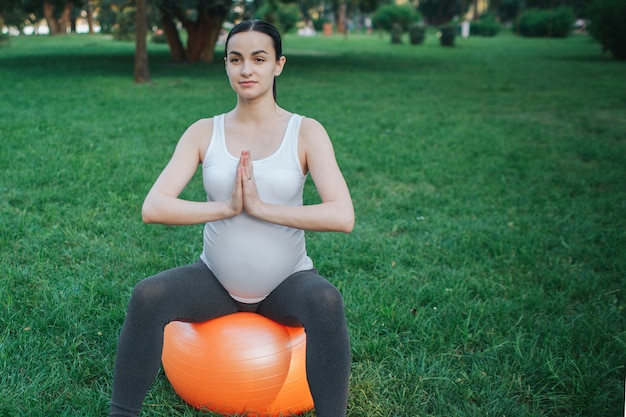 Concentré de jeune femme enceinte s'asseoir sur une boule orange fintess dans le parc. Elle se tient la main et médite.