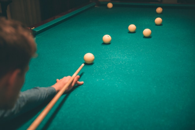 Concentré de jeune homme jouant. Il tient la queue de billard et vise à casser la balle sur le lit de la table. Il est seul dans la chambre.