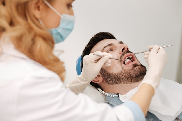 Concentré tendu bel homme couché dans la chaise de dentistes et tenant sa bouche ouverte pendant qu'elle examine sa santé dentaire