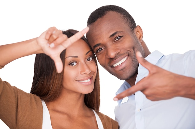 Concentrez-vous sur l'amour. Beau jeune couple africain regardant à travers un cadre de doigt et souriant en se tenant debout isolé sur blanc