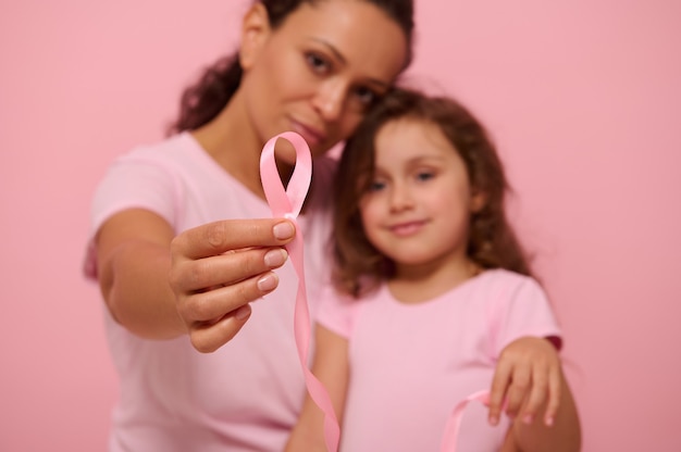 Concentrez-vous sur un long ruban de satin rose avec une extrémité sans fin entre les mains de deux générations floues de jolies femmes, maman et fille, exprimant leur soutien et leur solidarité aux patients atteints de cancer et aux survivants