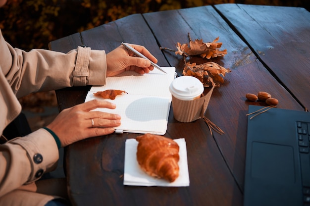 Concentrez-vous sur une main féminine tenant un stylo en argent et écrivant la liste à faire dans le bloc-notes, assis à une table en bois avec des feuilles de chêne sèches tombées, une tasse de papier à emporter et un croissant cuit au four. Image automne gros plan