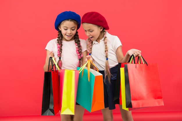 Concept d'accro du shopping. Signes que vous êtes accro au shopping. Les écolières mignonnes des enfants tiennent des sacs à provisions en tas. Élèves d'enfants satisfaits en faisant du shopping sur fond rouge. Obsédé par les centres commerciaux et les centres de vêtements.