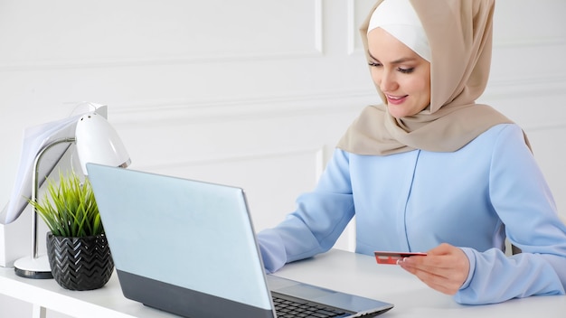Concept D'achat En Ligne. Portrait De Jeune Femme Musulmane En Hijab Beige Et Robe Bleue Traditionnelle Achète En Ligne Avec Une Carte De Crédit Et Un Ordinateur Portable.